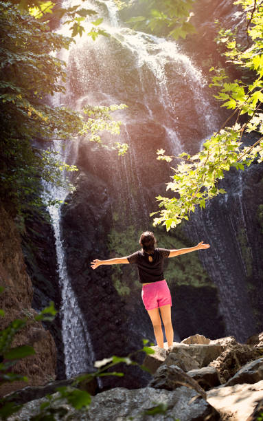 istock Woman standing by waterfall with her hands raised 829663938