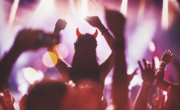 Cheering crowd at a concert. Closeup rear view of large group of people enjoying an open air concert on a summer night. There are a lot of hands raised. Blurry stage lights in background. An unrecognizable woman wearing glowing devil's horns is in focus as a silhouette. devil costume stock pictures, royalty-free photos & images