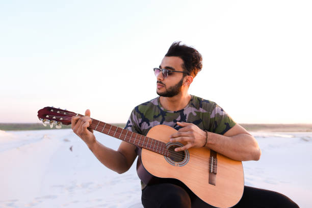 istock Handsome Arabian young man adjusts guitar, sitting on hill in mi 829750196