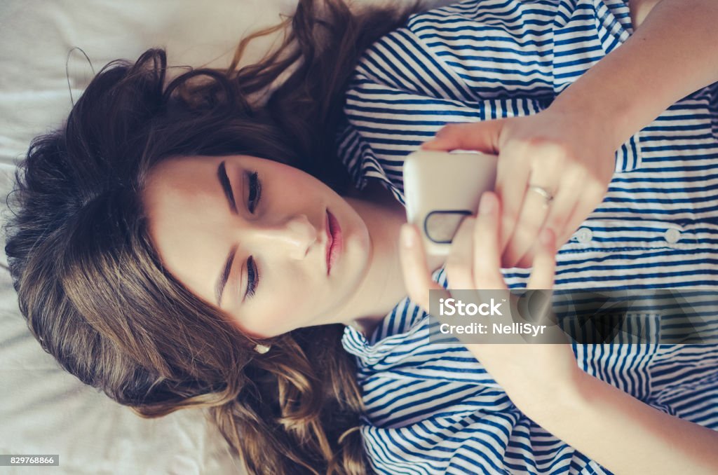 Closeup of pretty teenage girl lying in bed an looking at her mobile phone Closeup of sad teenage girl lying in bed using her mobile. Young pretty woman with bored expression looking at message on her cell phone, reading or waiting for call. Text Messaging Stock Photo