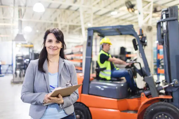 Female warehouse manager holding clipboard with a forklift loader in background