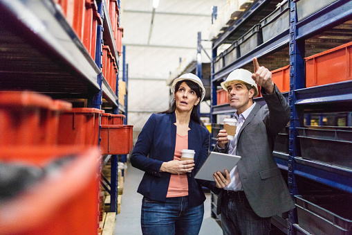 Manager with foremen controlling stock in warehouse. Businessman discussing with female worker in warehouse holding coffee and digital tablet.