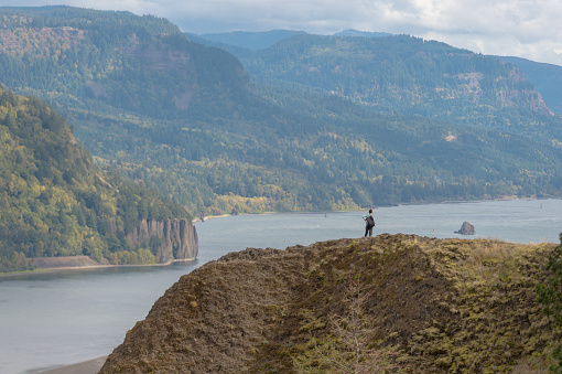 Far in the distance, a young solo female hiker stands atop a rock overlooking a mighty river. A mountain range rises up on the other side. The view is magnificent.