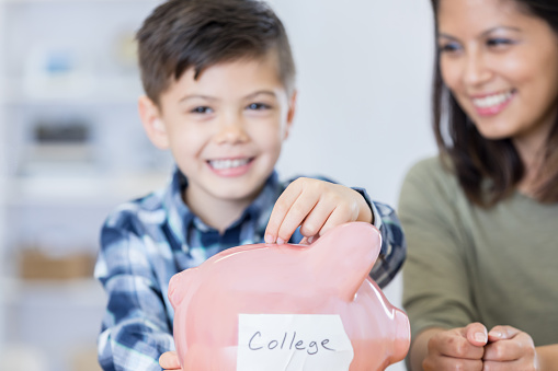 Little boy places coins in a piggy bank. The bank has 'College' written on the side. His mom teaching him about saving his money for college.