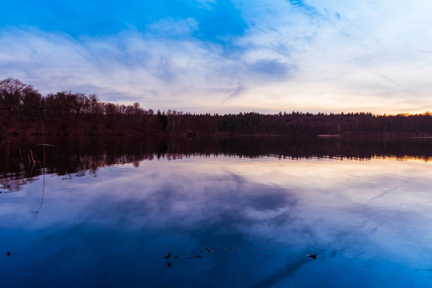 Calm rural lake reflecting an early sunset at dusk with the glow of the setting sun over the horizon and silhouetted trees on the shore reflected in the water