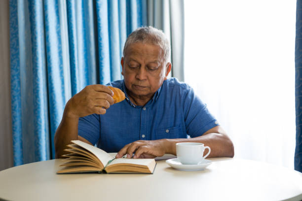 特別養護老人ホームで本を読んでいる間年配の男性が食べるクロワッサン - senior adult nursing home eating home interior ストックフォトと画像