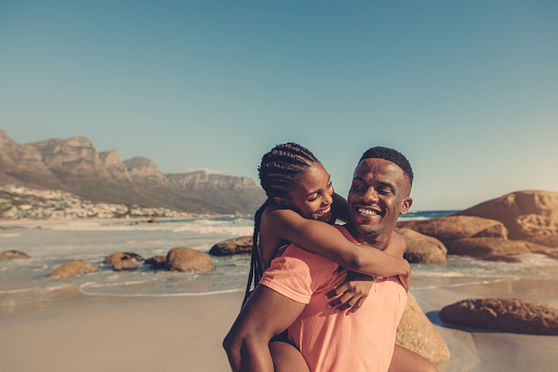 Handsome african man giving piggyback ride to his smiling girlfriend at the beach. Couple enjoying themselves at the seashore.