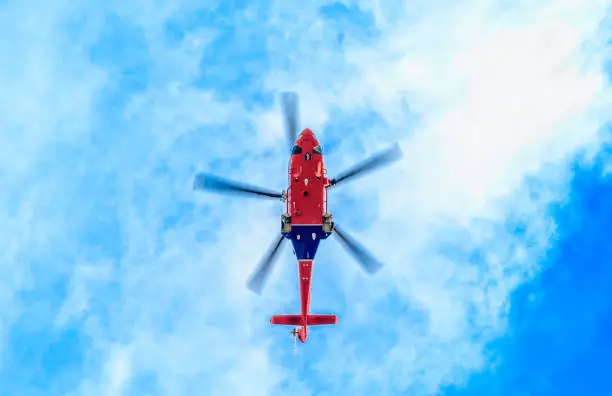 Looking up underneath a blue and red helicopter against a blue sky with light clouds