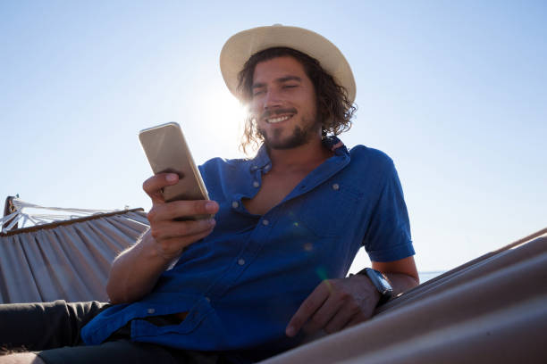 homme souriant, à l’aide de téléphone portable tout en vous relaxant sur le hamac sur la plage - hamac telephone homme photos et images de collection