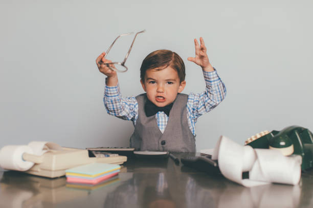 A little retro business boy dressed in bow tie, eyeglasses and vest answers the telephone at his business office desk. He is frustrated and angry at the chaotic situation at work and is overworked. He is holding up his eyeglasses in anger.