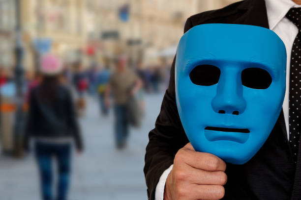 A Halloween theme close-up portrait of a young man holding up a white empty mask with his hand covering his mouth or half of his face, as he looks at the camera.\n\nA moody horror portrait of a person with a confusing identity, expressing negative emotions of confusion, anxiety and fear.
