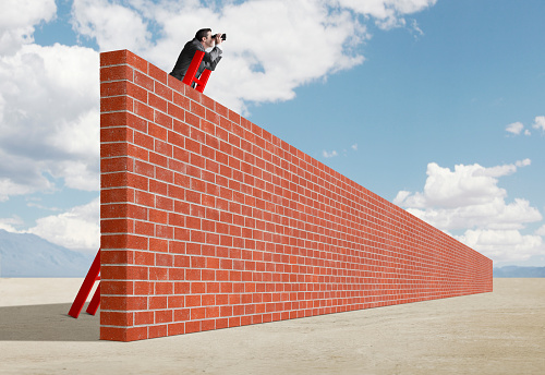 A businessman stands on a tall red ladder that leans against a brick wall that stands in the middle of a desert as he looks through a pair of binoculars as he tries to see what is in the distance.
