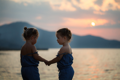 Identical twin girls standing on the sea shore holding hands with a beautiful landscape and sunset behind, Greece, Thassos