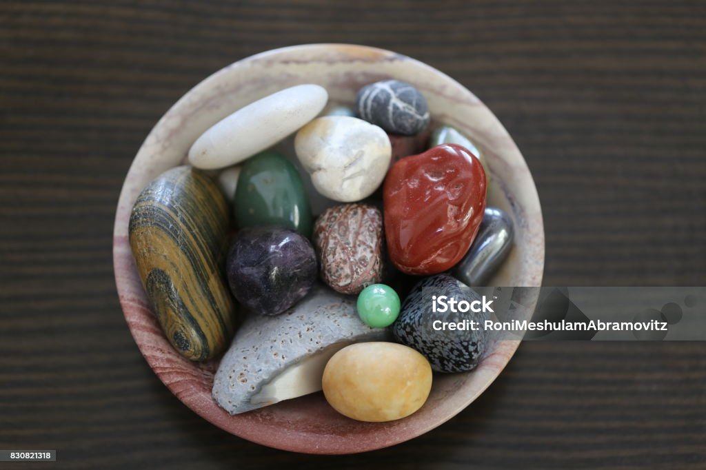 Colorful Semi Precious Stones. Assorted mix of smooth semi polished and waxed pebbles in a carved pink stone bowl on a wooden surface table. Collection of round natural semiprecious tumbled river stones and pebbles in variety of colors. Agate Stock Photo
