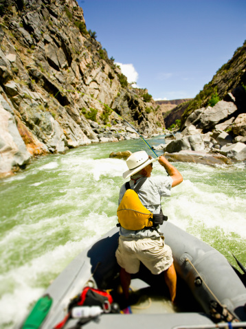 Late-50s man rafting and fly fishing on the Gunnison Gorge on the South Fork of the Gunnison River, Montrose, Colorado. He is standing up in the raft through a section of rapids while fishing. This section of river goes through dramatic rock canyons and is one of the most scenic rivers in Colorado.