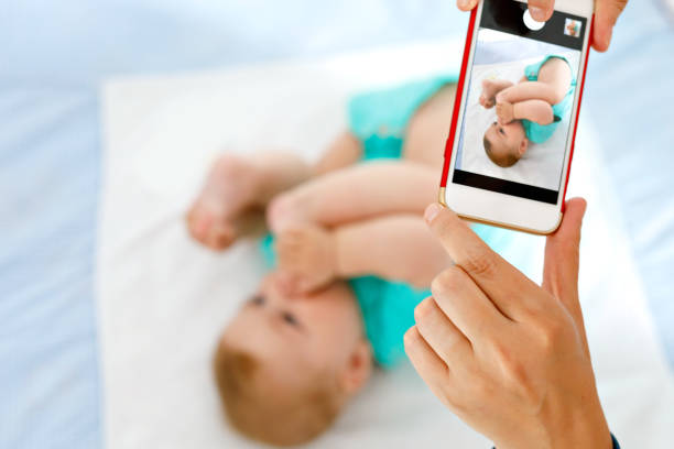 親のスマート フォンで赤ちゃんの写真を撮影。口の中に足を取って愛らしい新生子供 - 写真を撮る 写真 ストックフォトと画像