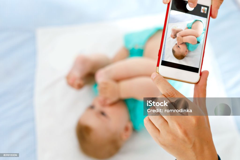 Parent taking photo of a baby with smartphone. Adorable newborn child taking foot in mouth Parent taking photo of a baby with smartphone. Adorable newborn child taking foot in mouth. sucking feet. Digital family memories Photographic Print Stock Photo