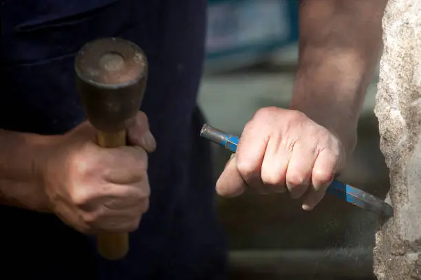 Close-up of craftsman's hands carving stone holding chisel, hitting with hammer, during a traditional public stone -carving festival, outdoors, sunlight lighting up chisel hand, spanish culture.