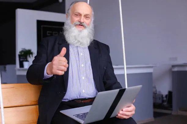 Old man holds laptop in hands and emotionally tells employees story or discusses at expense of questions in work, learns to understand programs and sits on suspended wooden bench on cables in comfortable recreation area of modern company. Elderly man with long gray beard of European appearance dressed in black classic suit and white shirt in blue stripes. Concept of elderly businessman in office, combining work during pension, training and assistance in mastering modern technology, lunch break after work.
