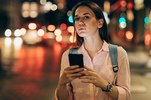 Young woman using a smart phone and waiting for her taxi in the city at night.