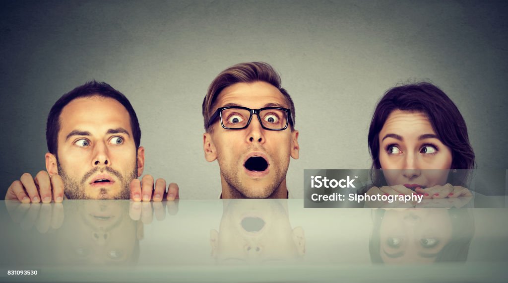 Scared young people two men and a woman hiding peeking form under the table Below Stock Photo