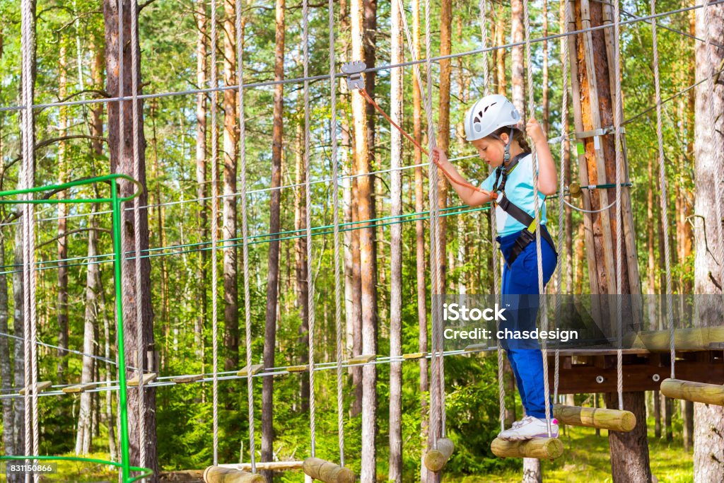 Girl on hinged trail in extreme rope Park Teenage girl goes on hinged trail in extreme rope Park in summer forest. High-altitude climbing training of child on adventure track, equipped with safety straps and protective helmet. Estonian summer Adventure Stock Photo