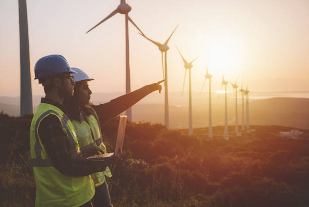 Young maintenance engineer team working in wind turbine farm at sunset Young electrical engineer woman and business man standing in front of wind turbines checking and working about technical problems and writes the results of measurements with laptop pc in wind power plant electric energy station. xxxl size taken with canon 5d mIV turbine stock pictures, royalty-free photos & images