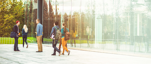 Group of students walking and standing in front of a modern glass university building, copy space.