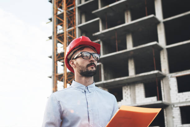 istock Young and modern engineer builder holds folder with drawings on background of building construction. Architect in helmet on construction site 831213314