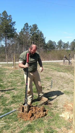 Muscular middle aged farmer digging post-hole for fence near hard packed dirt driveway.