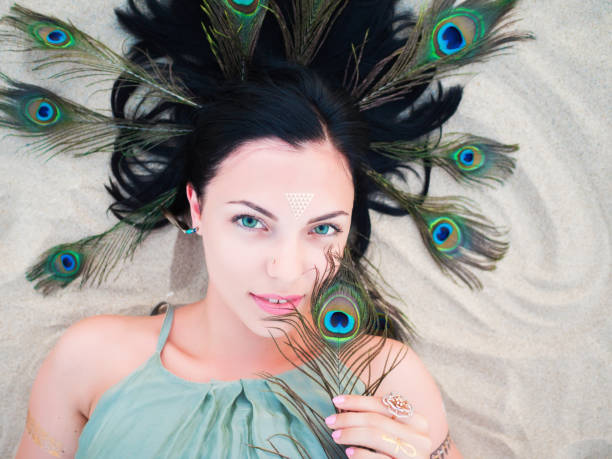 アウトドアファッションの髪で孔雀の羽と横になっているフラッシュの入れ墨をしたビーチで美しいブルネットの女性の肖像画。ジプシー自由奔放に生きるスタイル、緑のドレスの女 - ostrich feather animal head concepts ストックフォトと画像