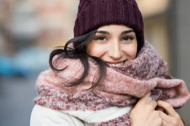 Closeup face of a young happy woman enjoying winter wearing scarf and cap. Smiling girl in a colorful shawl looking at camera. Latin woman with knitted bordeaux hat and woolen scarf."r