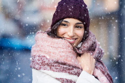 Smiling woman wearing wool bonnet and scarf in a winter cold day. Happy girl in a sweater outdoor in snow. Beautiful smiling woman with hat and scarf outdoor looking away.