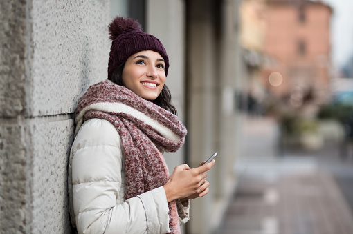 Pensive young woman holding smartphone and looking up. Happy smiling woman in winter with cap and woolen scarf thinking about the future. Latin girl standing on the street and looking away.