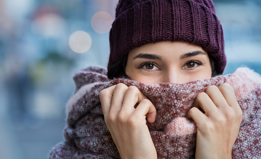 Winter portrait of young beautiful woman covering face with woolen scarf. Closeup of happy girl feeling cold outdoor in the city. Young woman holding scarf and looking at camera.\