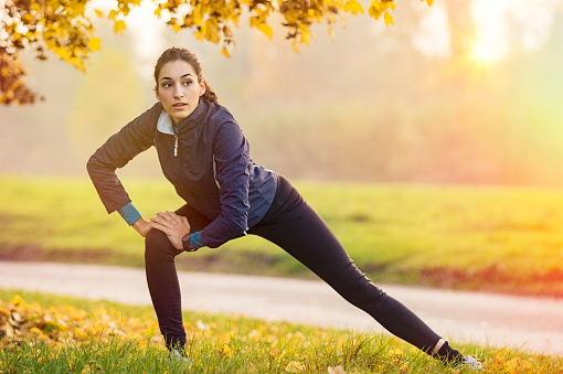 Young woman stretching and warming up at park during sunset. Attractive girl stretching before fitness in the autumn park. Beautiful sporty girl doing fitness outdoor during winter.