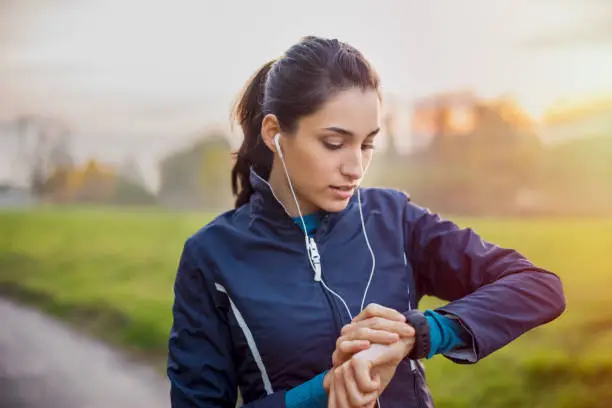 Young athlete listening to music during workout at park and adjusting smart watch. Young latin woman setting smartwatch before jogging in winter or autumn."r