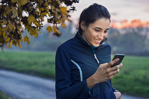 Young athlete looking at phone and smiling at park during sunset. Smiling young woman checking her smartphone while resting. Happy girl texting a phone message with a big smile on her face.