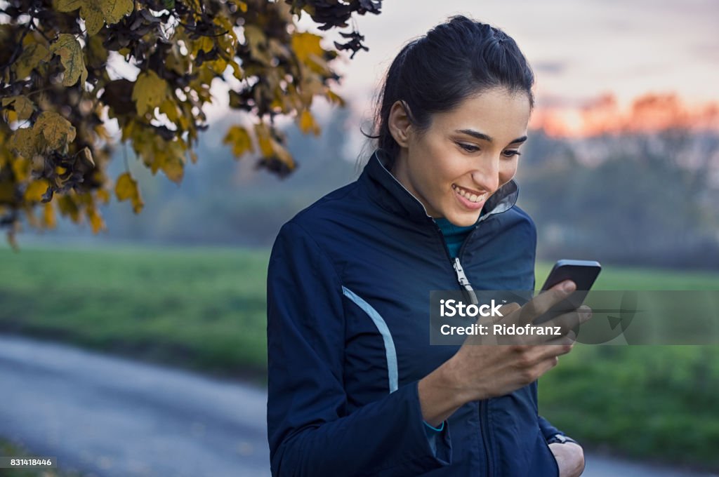 Sporty woman using phone Young athlete looking at phone and smiling at park during sunset. Smiling young woman checking her smartphone while resting. Happy girl texting a phone message with a big smile on her face. Using Phone Stock Photo