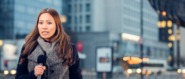 Female newsreader Front view of a female asian ethnicity newscaster wearing coat talking into microphone, blurred city buildings in background, copy space. newsreader stock pictures, royalty-free photos & images