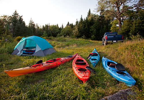 a wilderness campsite with a pickup truck, four colorful kayaks, and a pitched tent in the woods