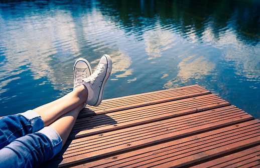Woman relaxes by the lake sitting on the edge of a wooden jetty , swing one's feet near the water surface. Sunny joyful summer day concept.