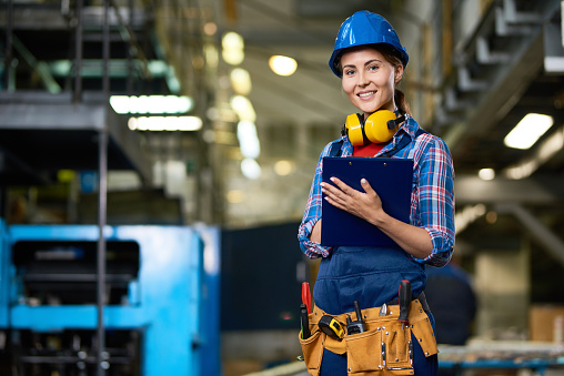 Portrait of female construction worker working in modern plant looking at camera, smiling filling in papers on clipboard