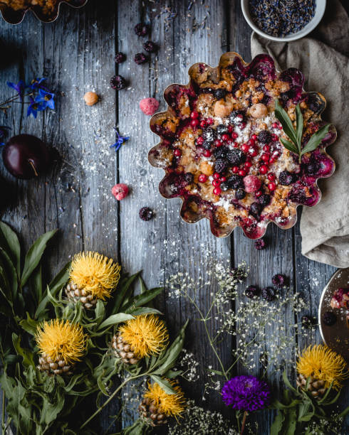 currant and raspberry pie with colorful flowers and plums on a wooden table in rustic style tarta de bayas surtidas en un metal forma con textiles, flores amarillas y púrpuras, plato pequeño con lavanda seca, ciruelas, plato con una rebanada de pastel y una cuchara sobre un fondo gris de la madera, la vista superior de la hornada