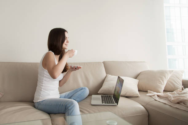 istock Young woman enjoying coffee on sofa with laptop, day off 831554182