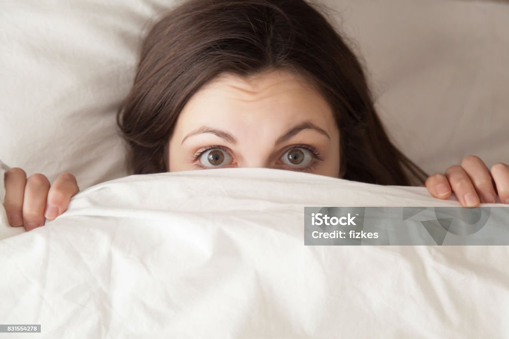 Funny surprised girl covering face with white blanket, headshot closeup Funny surprised girl covering half of face with white blanket, young scared woman hiding and peeking from duvet, afraid of night monsters, feels embarrassed, wide awake, head shot close up, top view Dreamlike Stock Photo