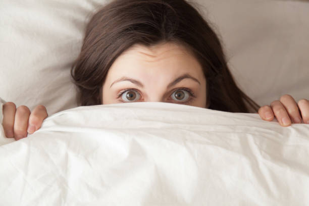 Funny surprised girl covering face with white blanket, headshot closeup Funny surprised girl covering half of face with white blanket, young scared woman hiding and peeking from duvet, afraid of night monsters, feels embarrassed, wide awake, head shot close up, top view horror waking up bed women stock pictures, royalty-free photos & images