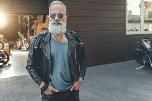 Severe aged biker in goggles is standing near open garage. He putting hands in pockets and glancing at camera. Portrait. Copy space