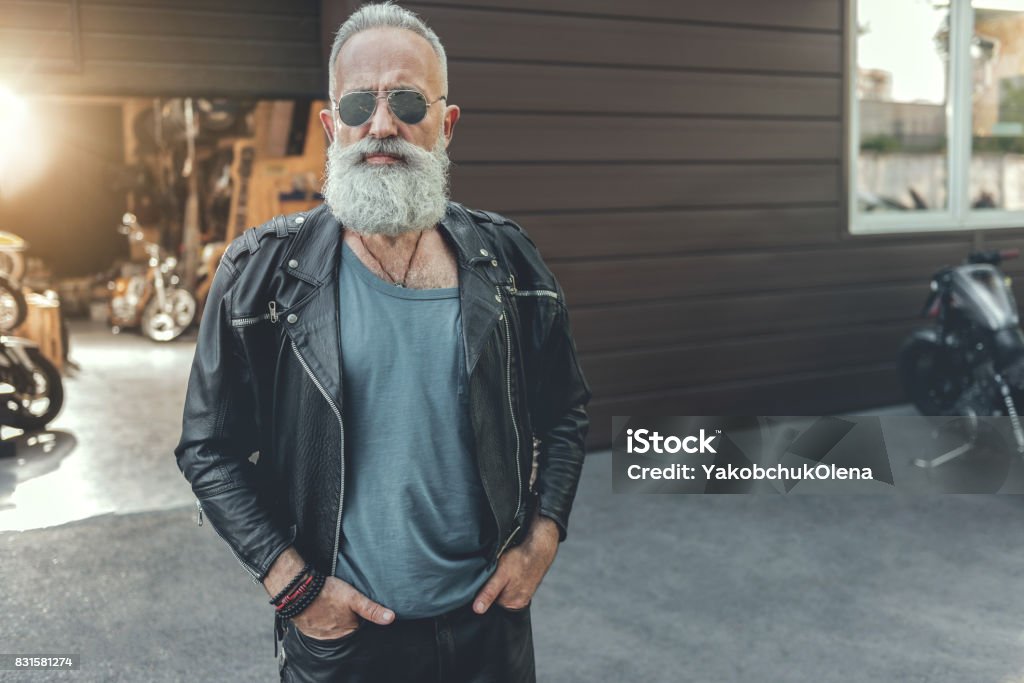 Serious bearded old man wearing leather Severe aged biker in goggles is standing near open garage. He putting hands in pockets and glancing at camera. Portrait. Copy space Biker Stock Photo