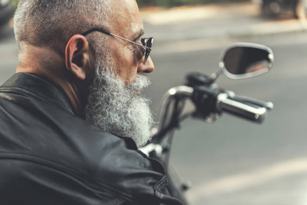 Old male person on motorbike Aged bearded biker in sunglasses is sitting at motorcycle and looking aside. He wearing jacket made of black leather. Focus on face. Profile serious monkey stock pictures, royalty-free photos & images
